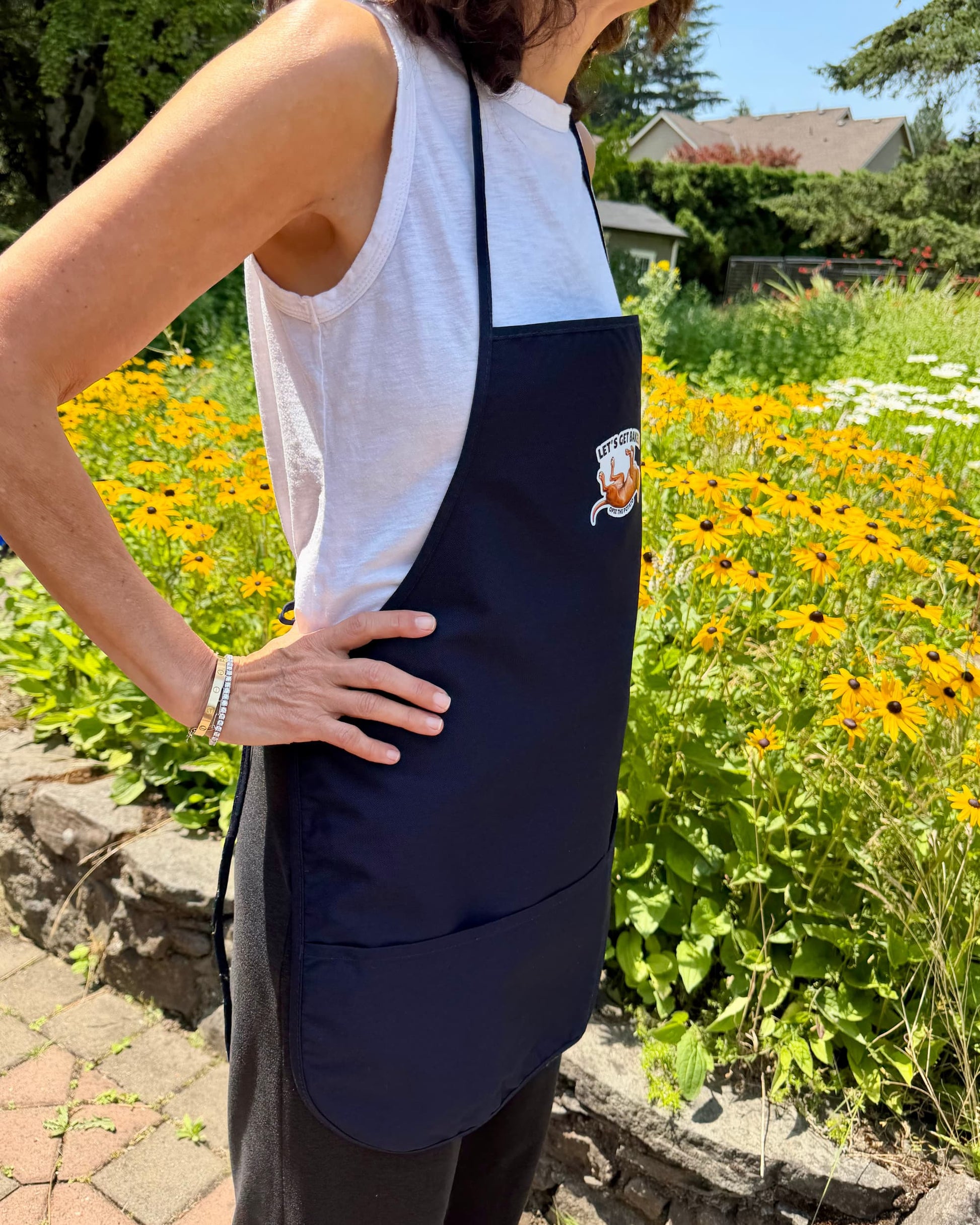 Opie Let's Get Baked apron worn by woman from side view in garden with yellow flowers, navy blue cooking apron with Opie the Potato graphic, official @justopiethings merch by Sly Dawg Studio