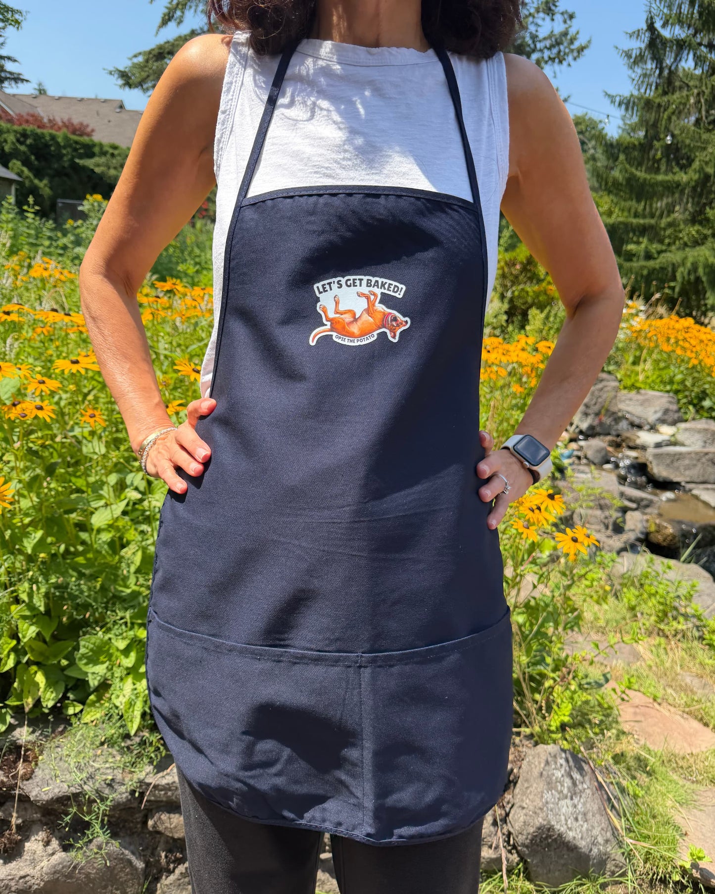 Opie Let's Get Baked apron worn by woman facing forward in garden, navy blue bib apron showing full Let's Get Baked pit bull graphic on chest with two front pockets, official @justopiethings merch by Sly Dawg Studio
