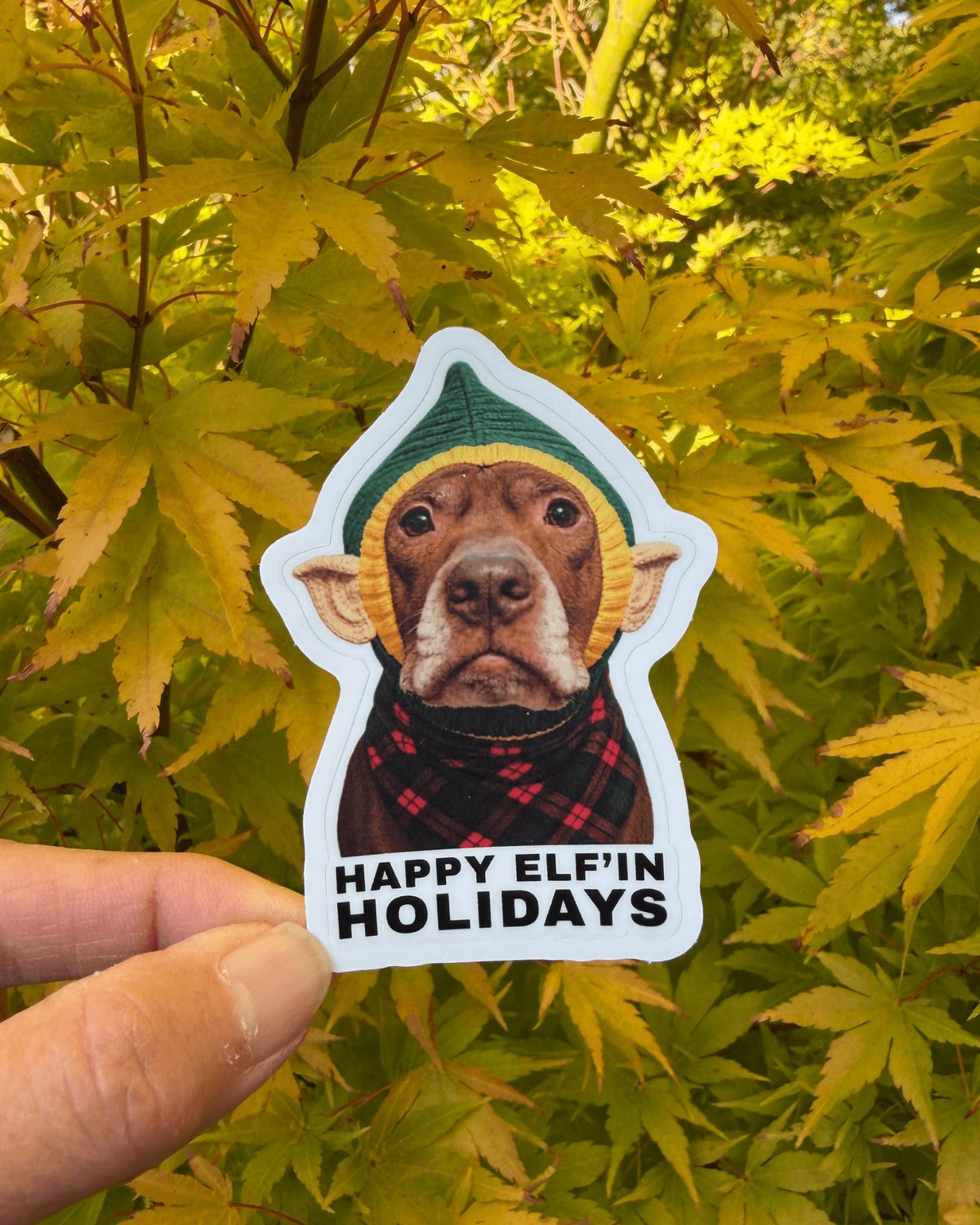Hand holding the Opie Christmas elf sticker in front of autumn leaves