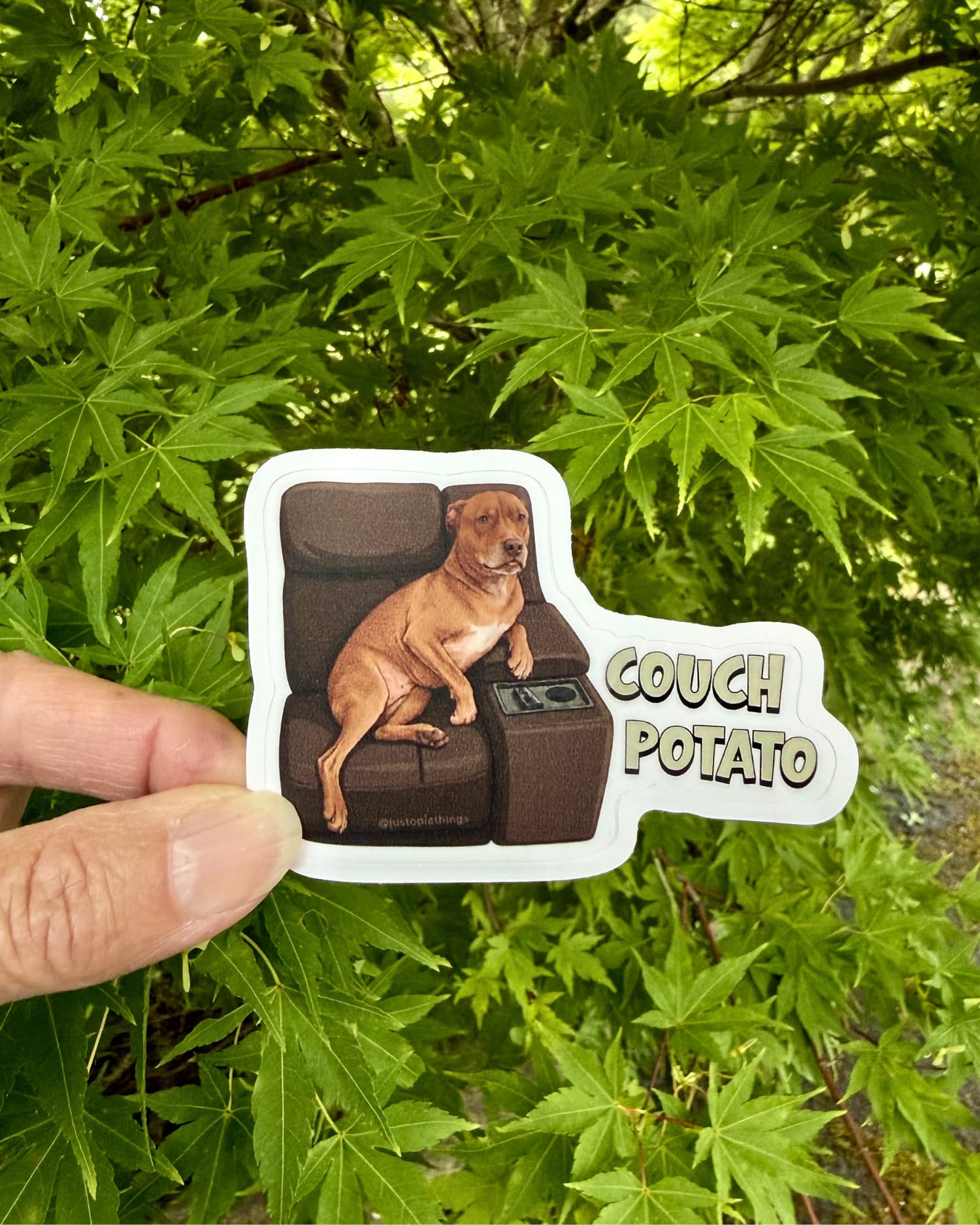 Hand holding Opie Couch Potato sticker outdoors against green Japanese maple leaves