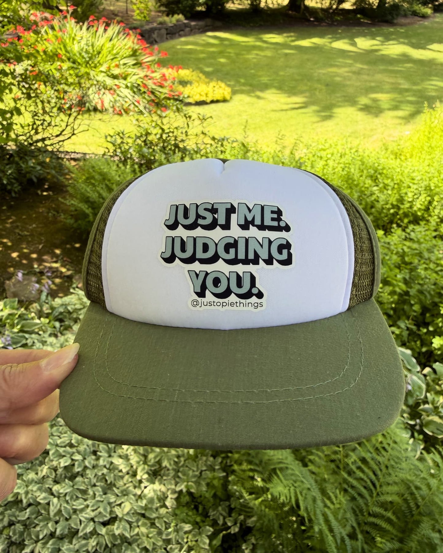 handheld in front of yard greenery: Opie Dog Hat front view, olive green and white with Just Me Judging You text and justopiethings handle.
