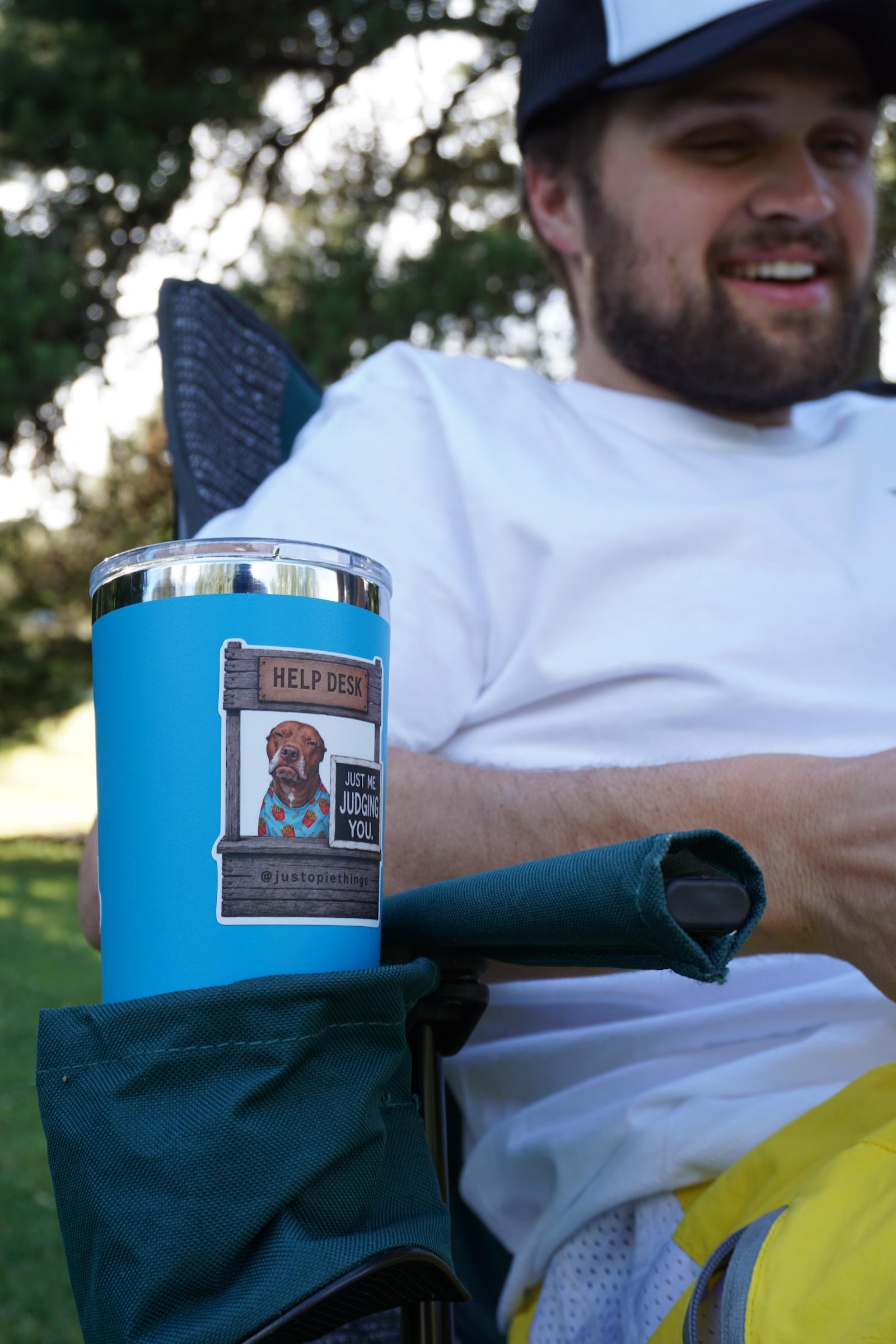 A bright blue insulated tumbler cup with a clear lid and a printed graphic Opie Help Desk Sticker on the front, held in a dark teal chair cup outdoors. The graphic is Opie the pit bull sitting at a help desk with a sign that says "Just me. Judging you.