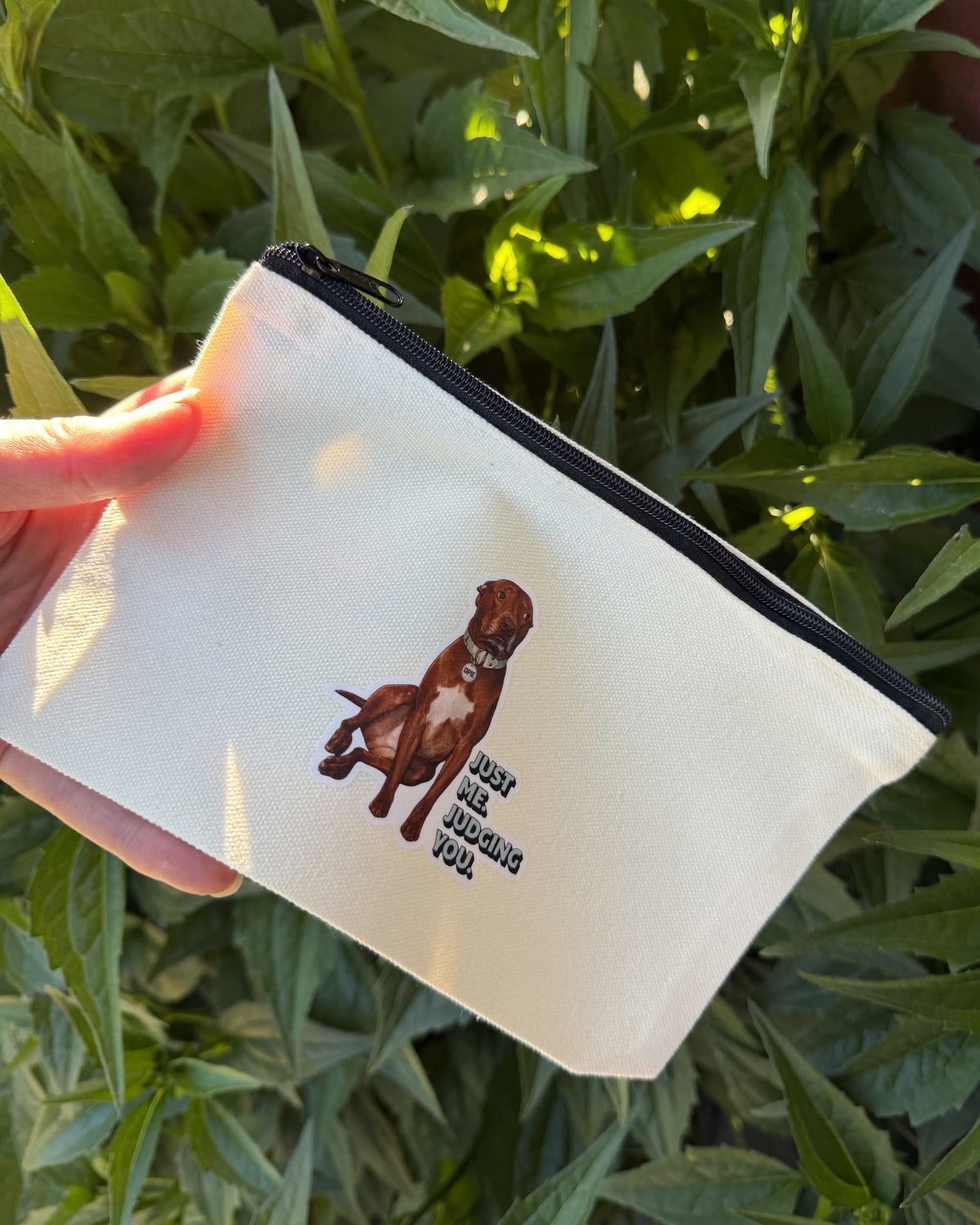 Opie zipper pouch. Canvas bag with top zipper and design of pit bull on front shown in front of leaves on a bush.