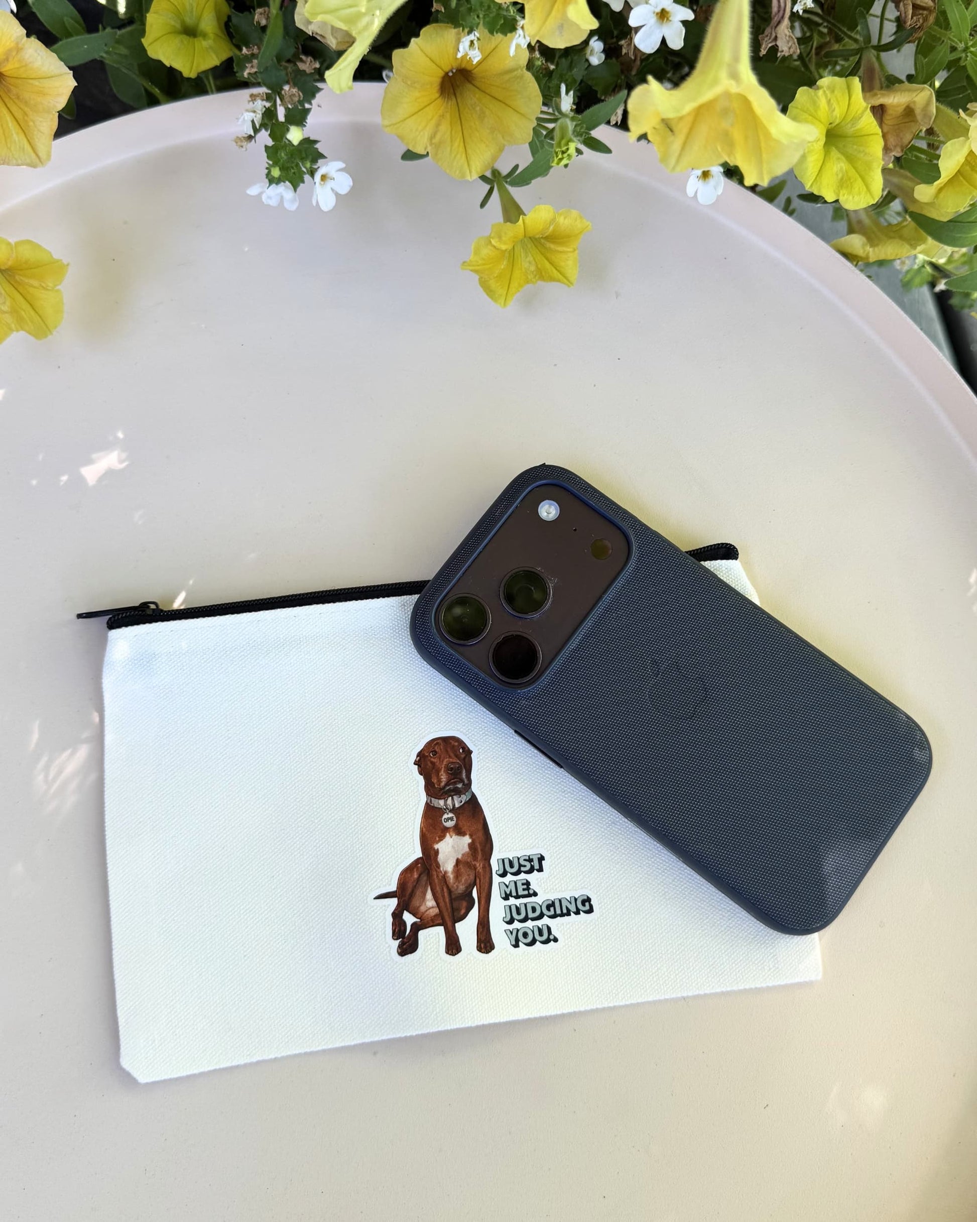 Ope zipper pouch shown on a pale yellow table. Natural canvas bag has black zipper and brown pit bull design on front with text "Just me. Judging you." A phone with navy case is on the bag to show size.