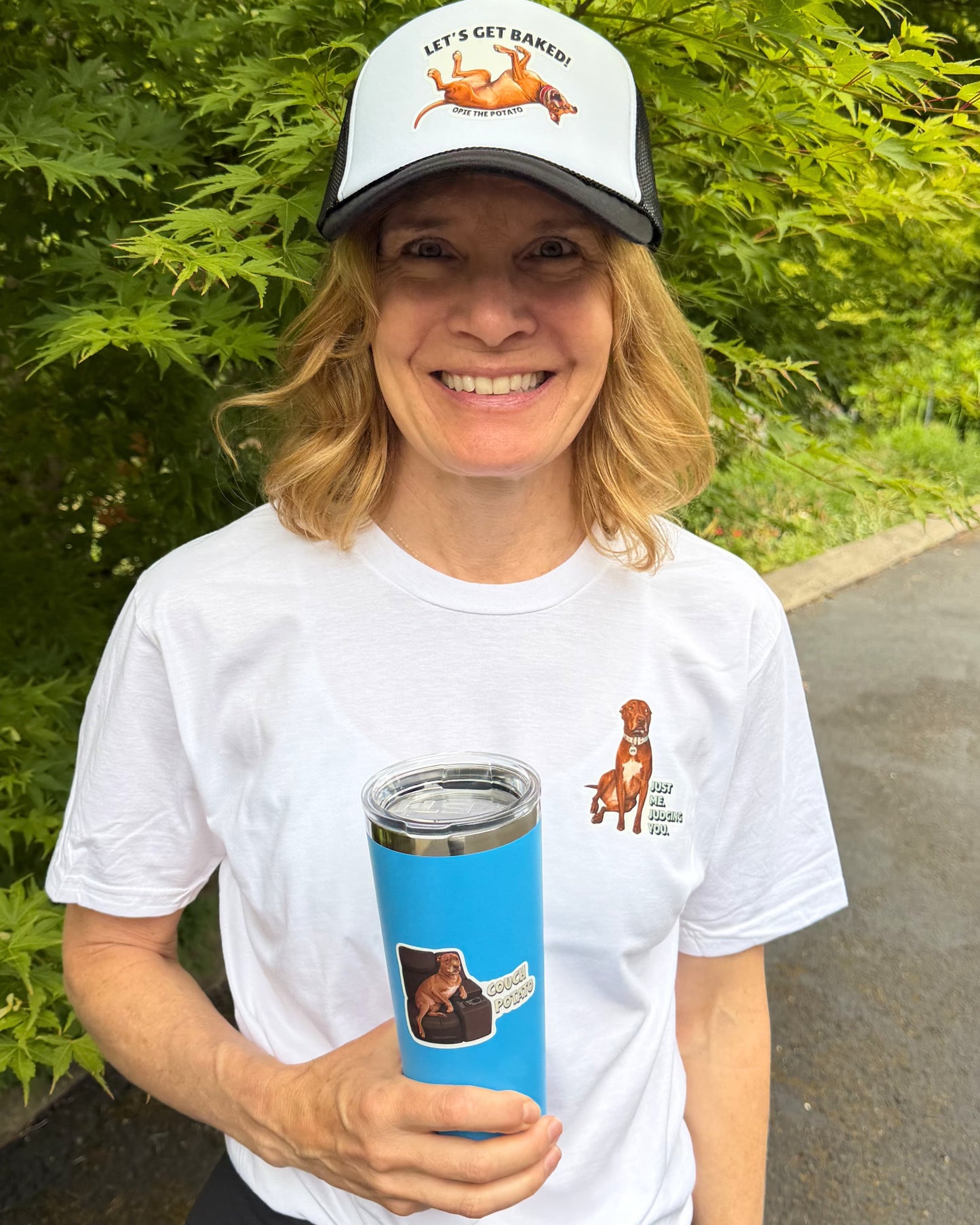 Woman smiling outdoors wearing white Opie just me judging you t-shirt and Let's Get Baked trucker hat holding a blue tumbler with Couch Potato sticker