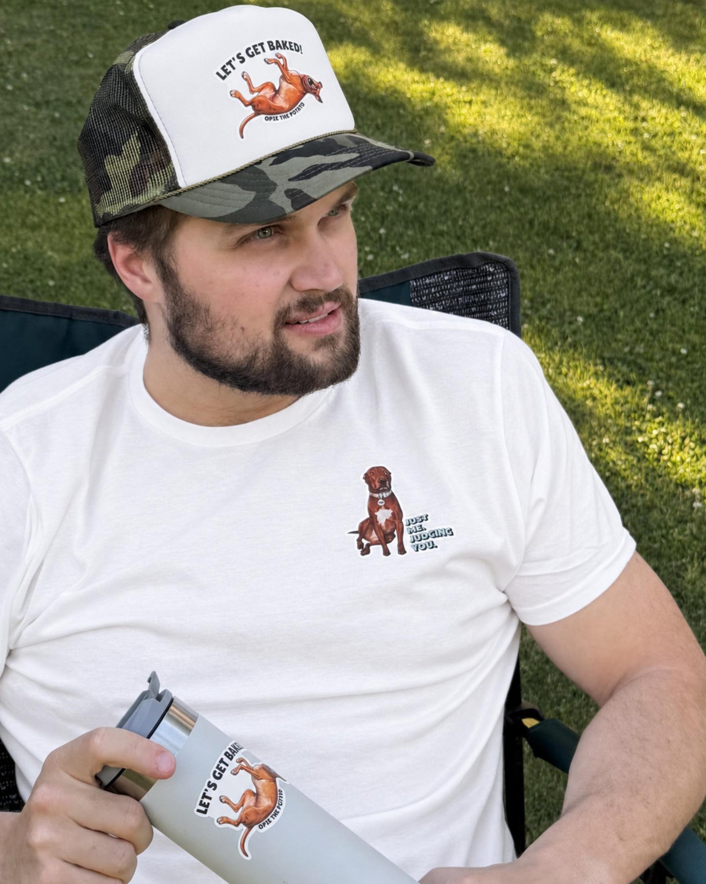 Man wearing white t-shirt with Opie the pit bull Just Me. Judging You. left chest design, seated outdoors in grass wearing Opie Let's Get Baked camo trucker hat and holding tumbler with Let's Get Baked sticker