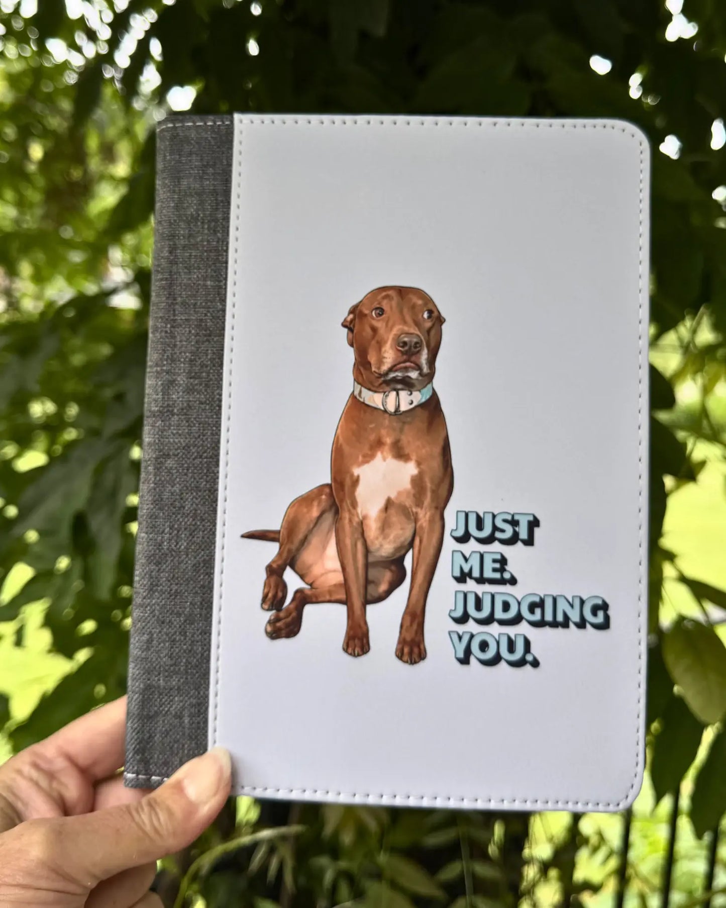 Opie reusable notepad portfolio held outdoors, white faux leather cover with Just Me. Judging You. pit bull design and charcoal fabric spine, official justopiethings merch by Sly Dawg Studio
