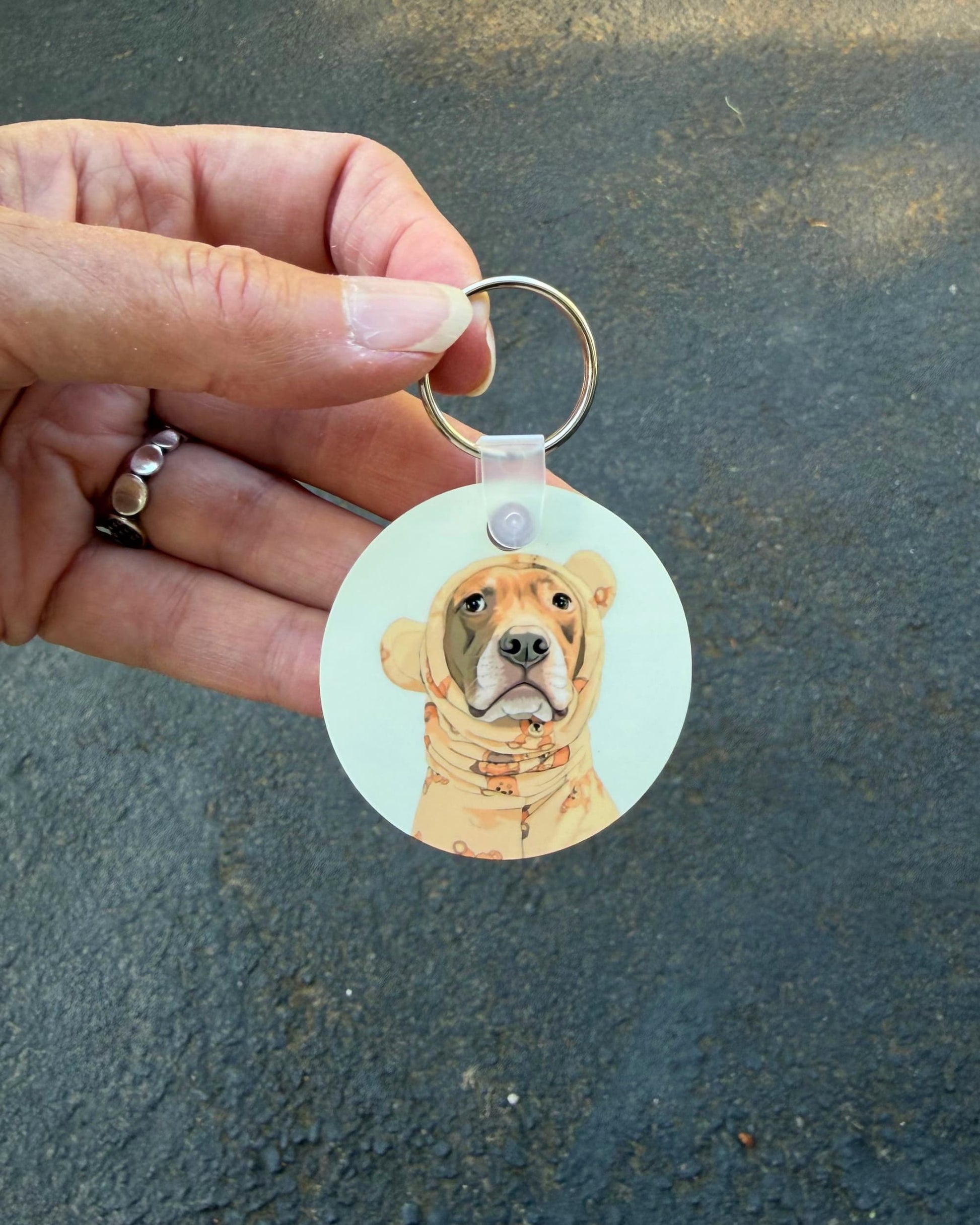 Opie keychain held between fingers outdoors, round aluminum pit bull keychain featuring Opie in teddy bear pajamas, official @justopiethings merch by Sly Dawg Studio