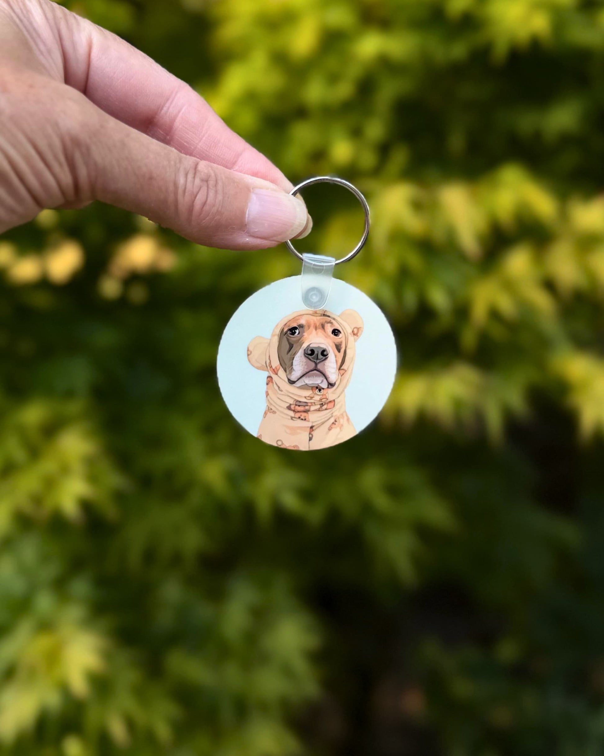 Opie keychain held up outdoors showing pit bull in teddy bear pajamas design on mint green background, official @justopiethings merch by Sly Dawg Studio