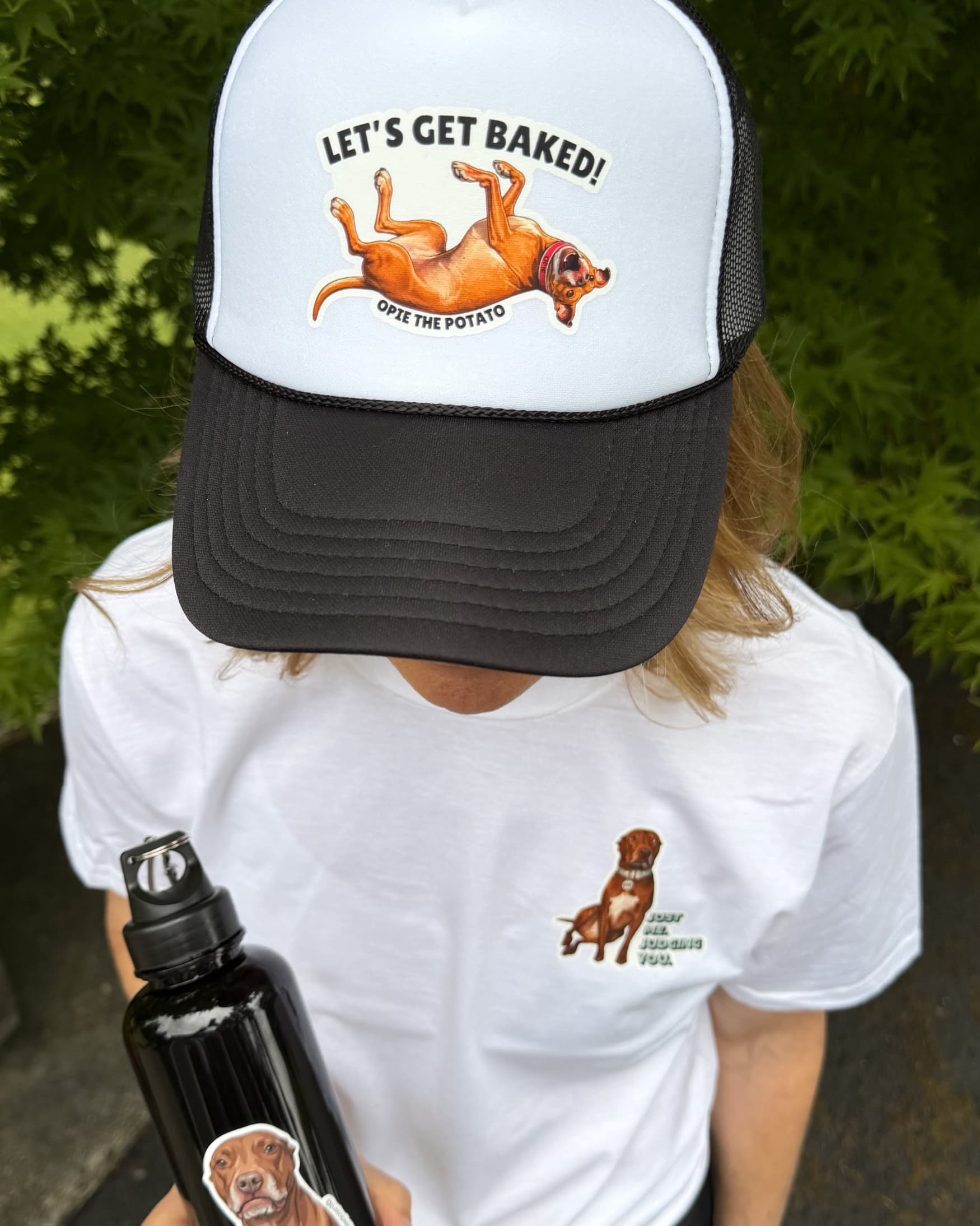 Person wearing Opie Lets Get Baked Trucker Hat for Humans in black outdoors, downward angle showing hat design