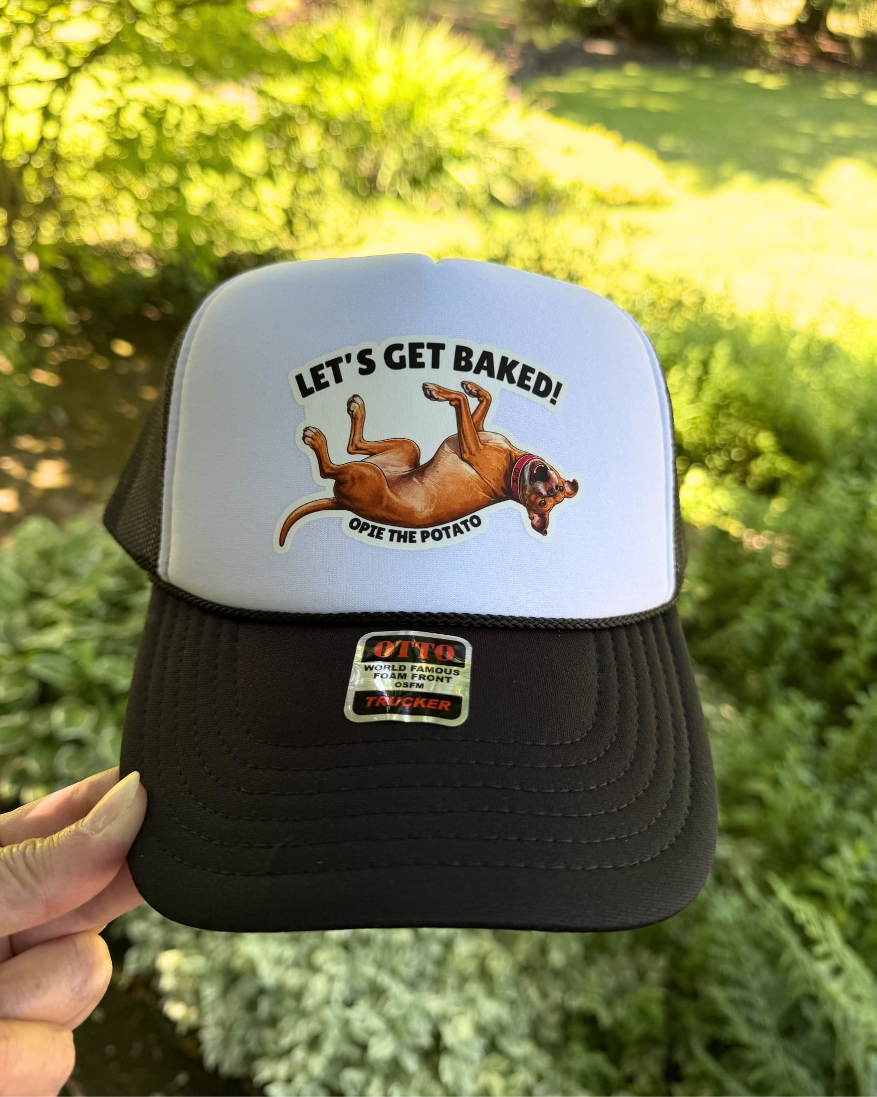 Person holding Opie Lets Get Baked Trucker Hat for Humans in camo outdoors, showing Opie the Potato Lets Get Baked design