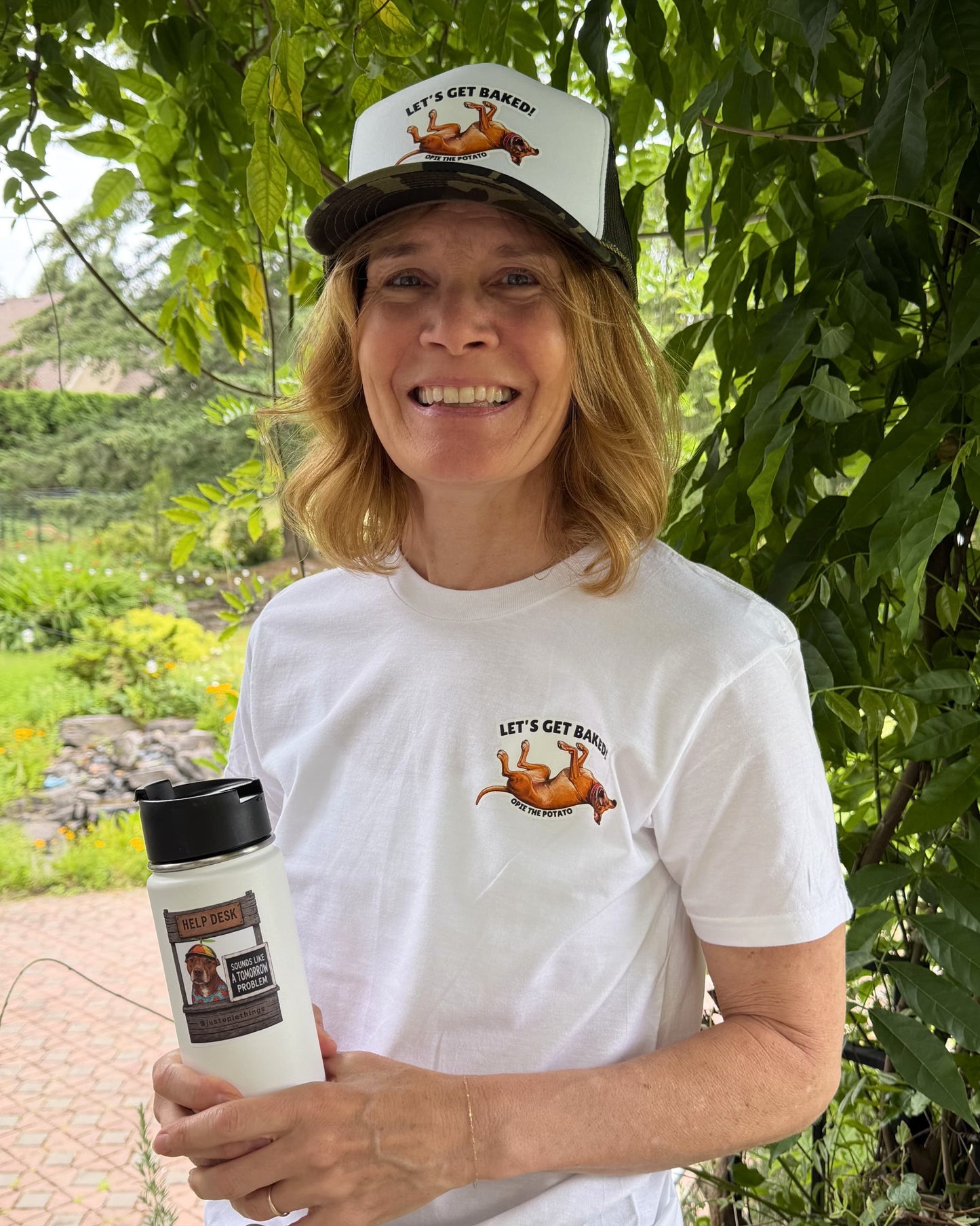 Woman smiling outdoors wearing white Opie lets get baked t-shirt and matching trucker hat holding a tumbler
