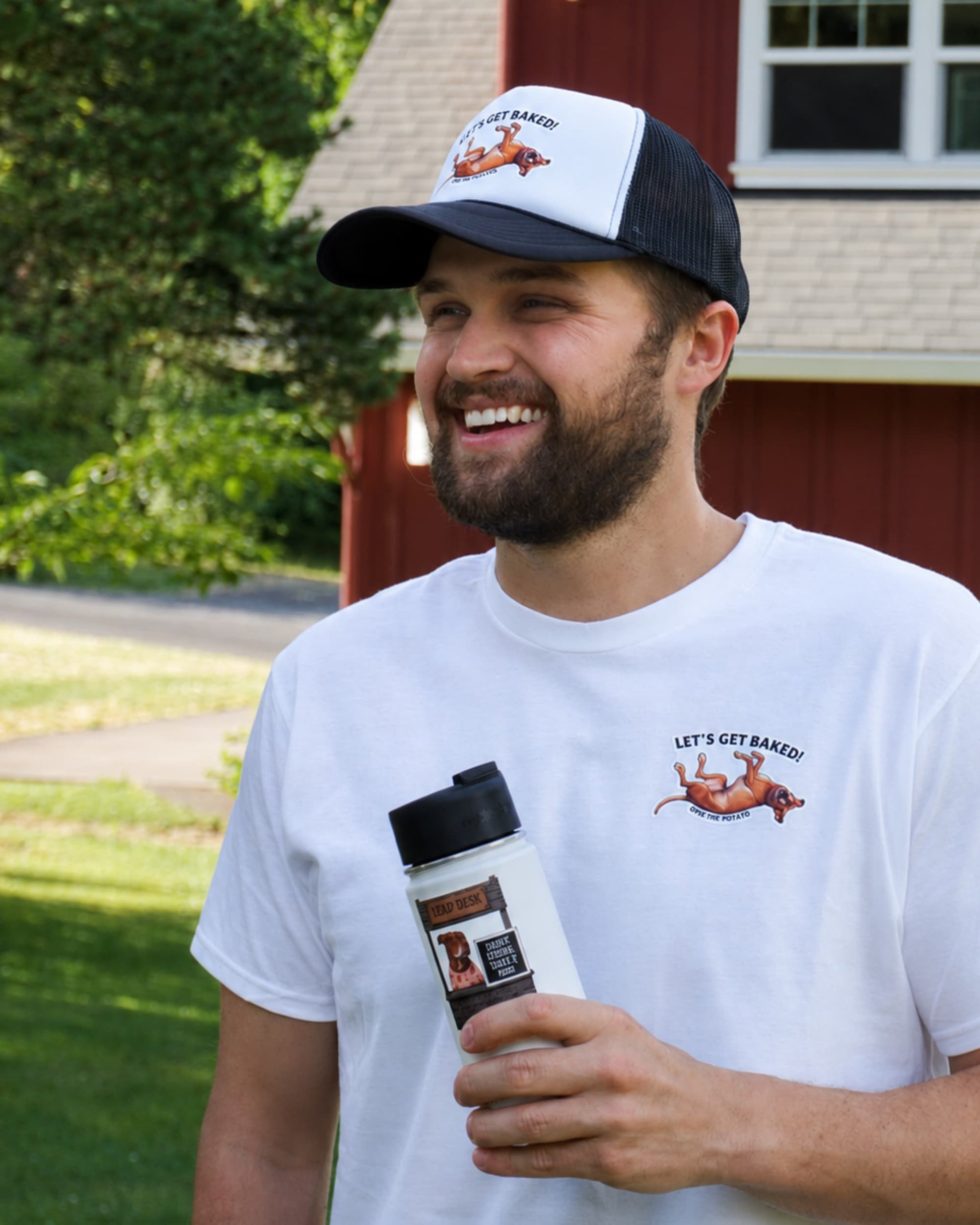 Opie let's get baked t-shirt: A smiling man outdoors wearing a white graphic Opie T-shirt and a black-and-white Opie trucker hat, holding a white insulated bottle. The shirt has a small dog graphic and the text 'LET'S GET BAKED!' on the left chest.