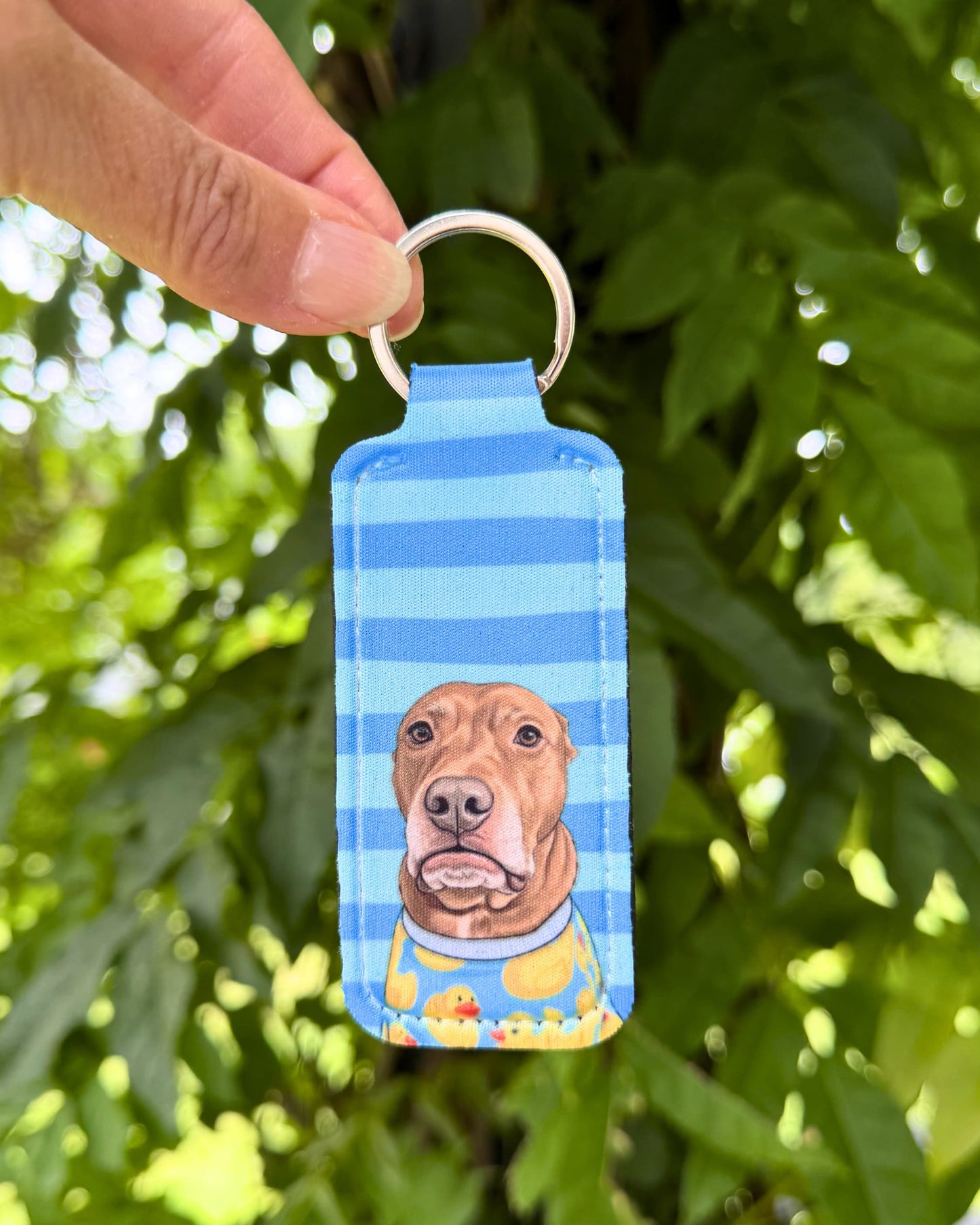 Opie lip balm holder keychain in blue stripe design held outdoors against green foliage, pit bull portrait on neoprene lip balm holder, official @justopiethings merch by Sly Dawg Studio