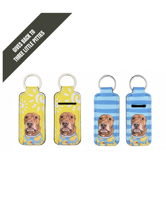 Opie lip balm holder keychain three pack showing yellow polka dot sun and blue stripe designs with pit bull portraits, Give Back product supporting Three Little Pitties Rescue, official @justopiethings merch by Sly Dawg Studio. Gray corner banner "Gives back to THREE LITTLE PITTIES"