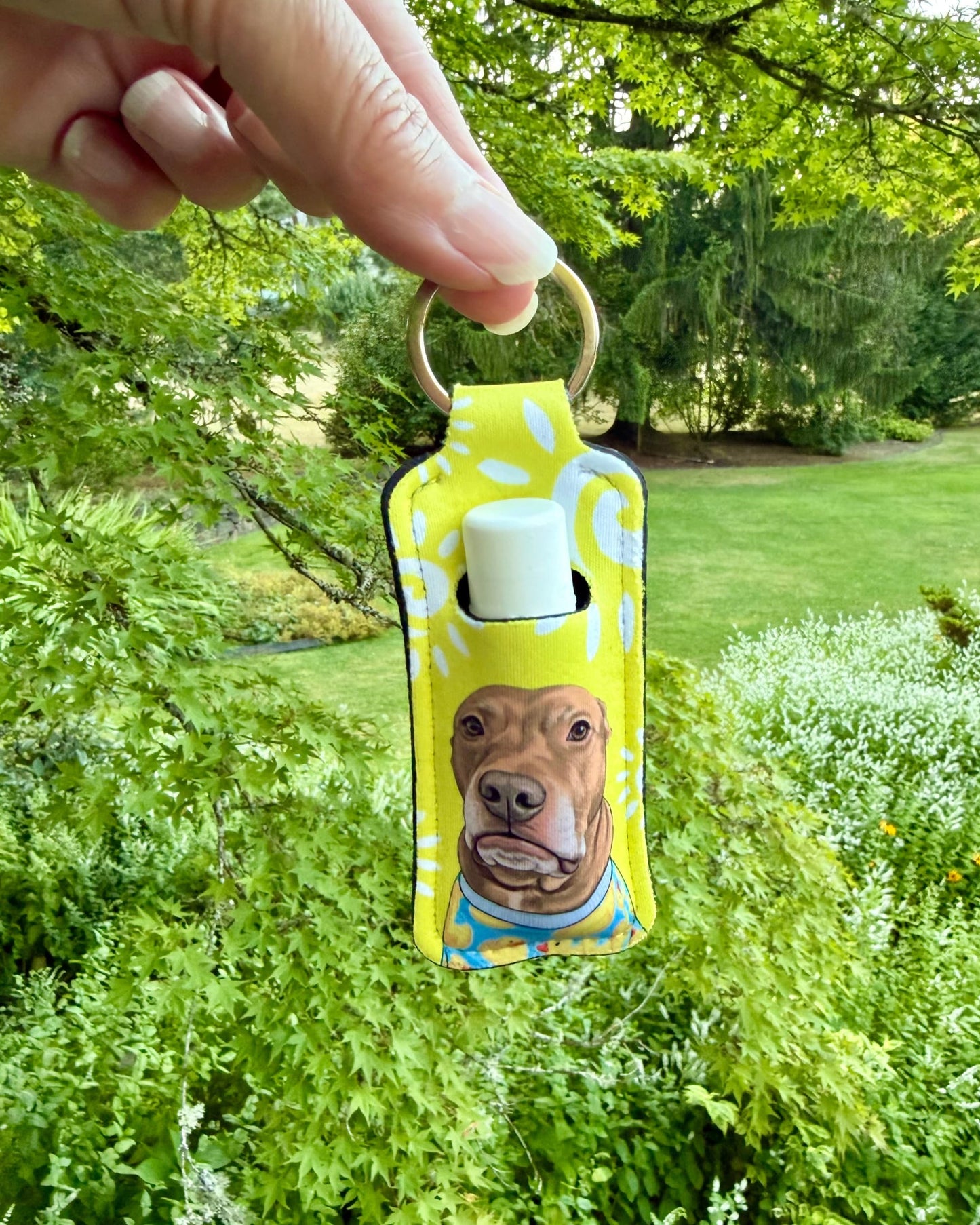 Opie lip balm holder keychain in yellow polka dot design held outdoors against green foliage, pit bull portrait on neoprene lip balm holder, official @justopiethings merch by Sly Dawg Studio