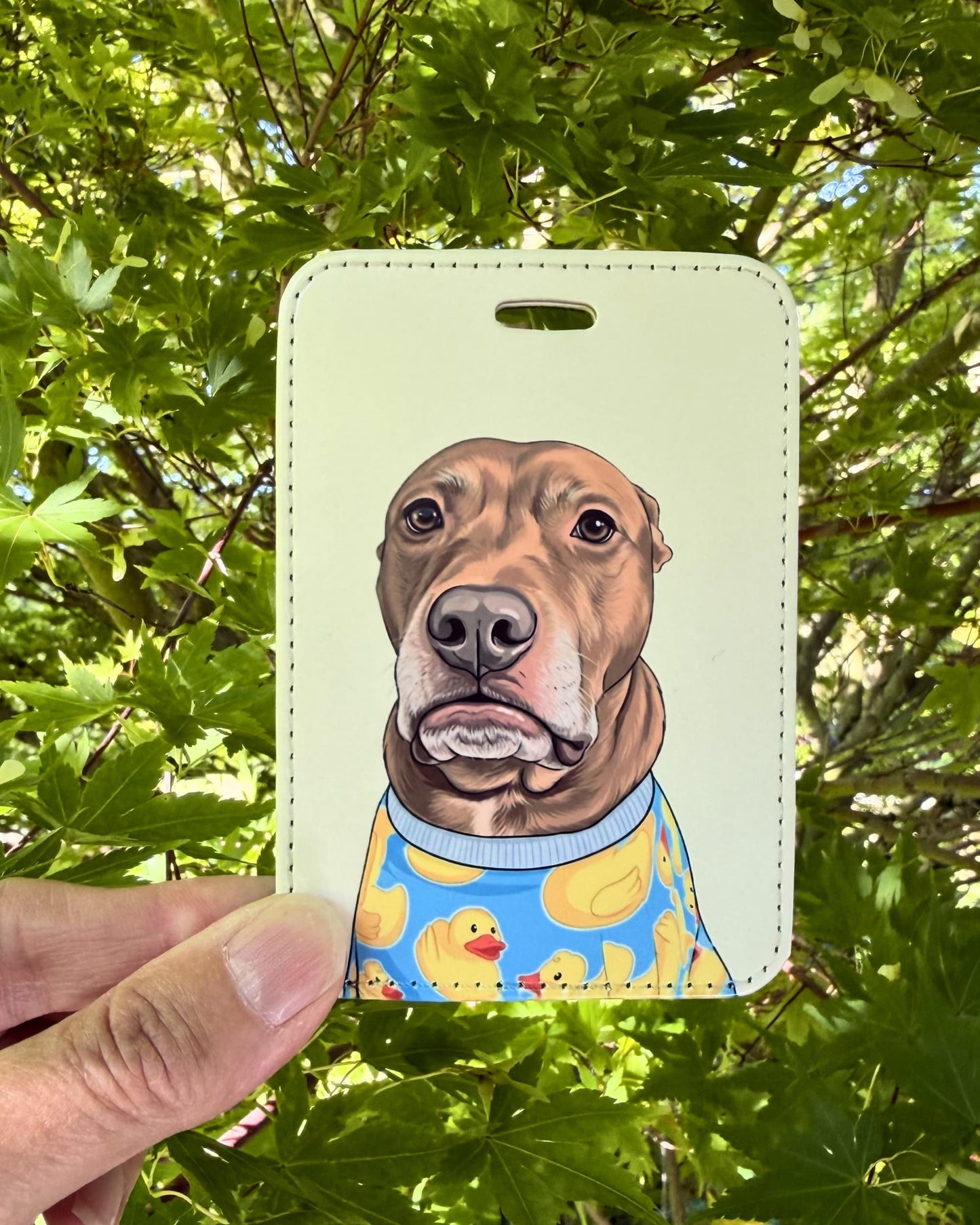 Hand holding Opie duck pajamas faux leather luggage tag outdoors against green leaves showing print quality and color.