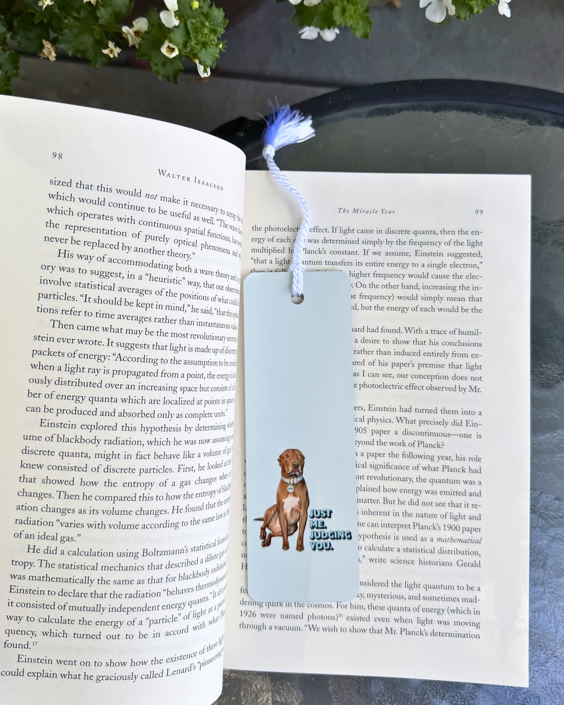 Opie Metal Bookmark, white metal bookmark with white tassel featuring Opie the pit bull sitting with Just Me Judging You text on light gray background. Shown inside a book on an outdoor table.