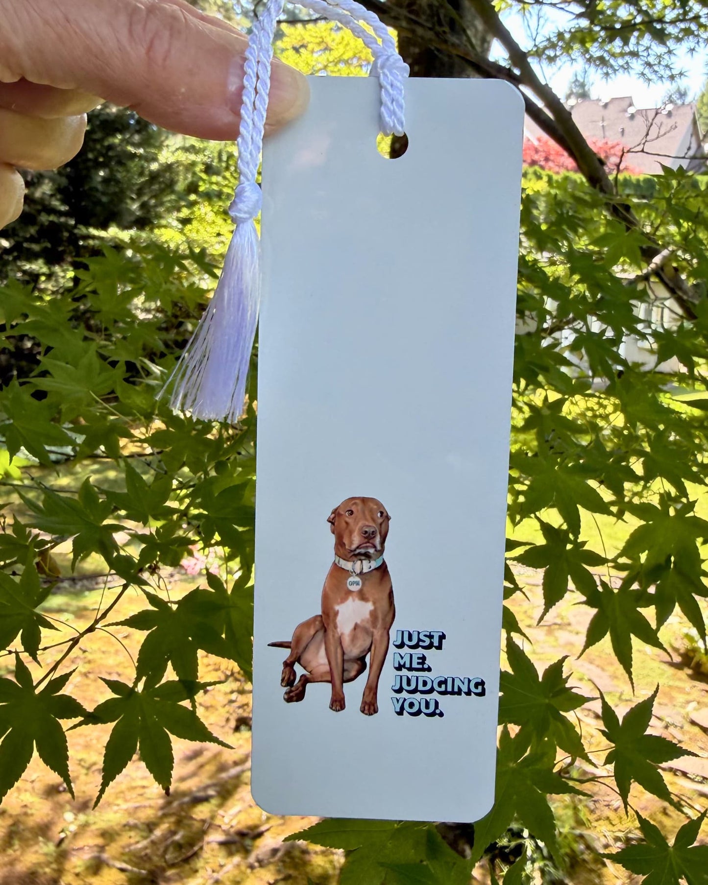 Opie Metal Bookmark held outdoors against Japanese maple tree, full length view with white tassel