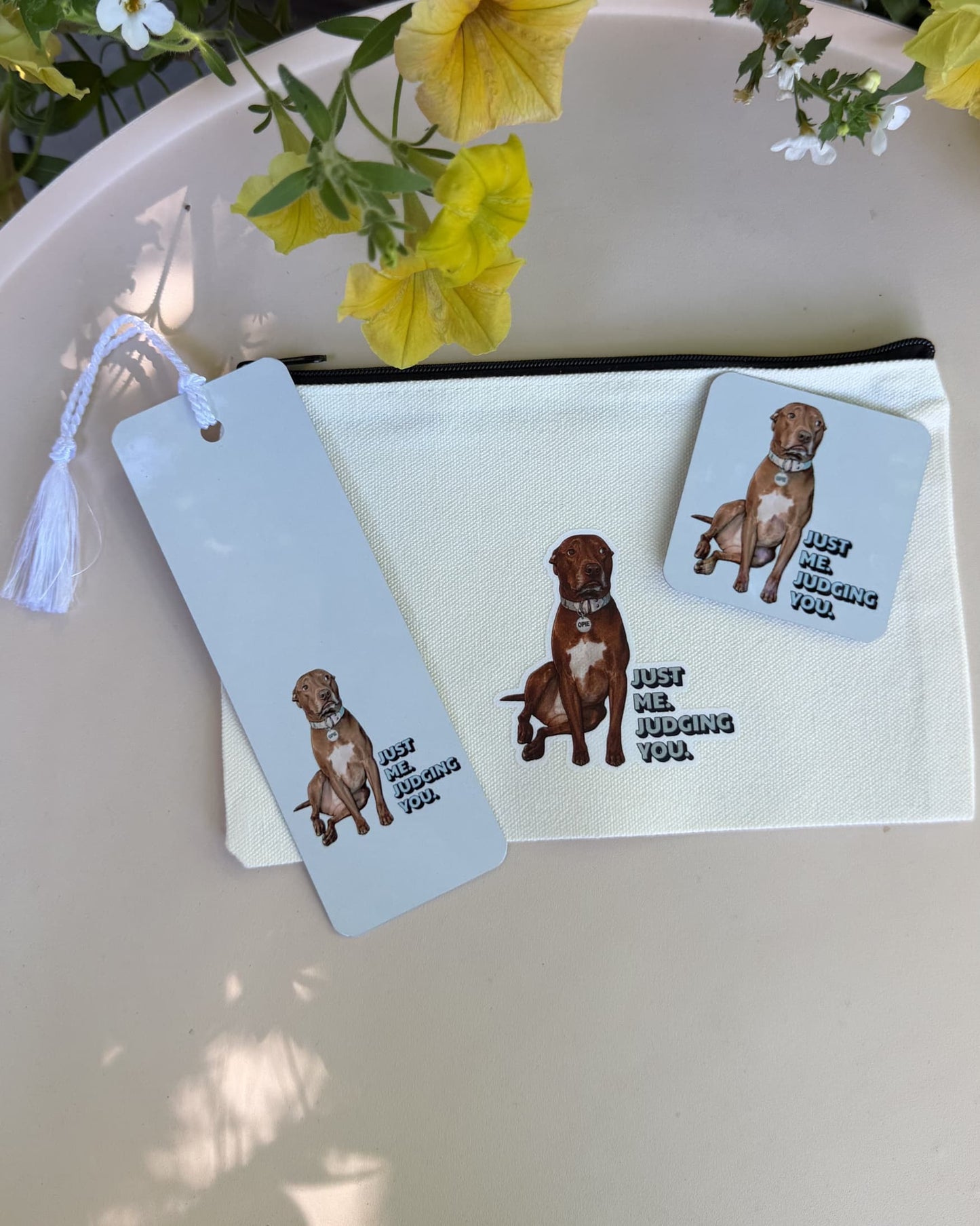 Opie Metal Bookmark shown with Opie zipper pouch and Opie magnet on white outdoor table with yellow petunias