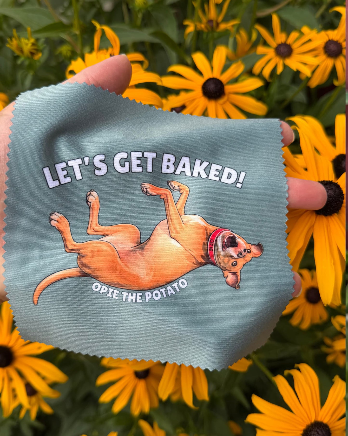 Opie the Potato microfiber cloth held in hand against yellow flowers, slate blue design with "Let's Get Baked!" text and rolling pit bull dog illustration