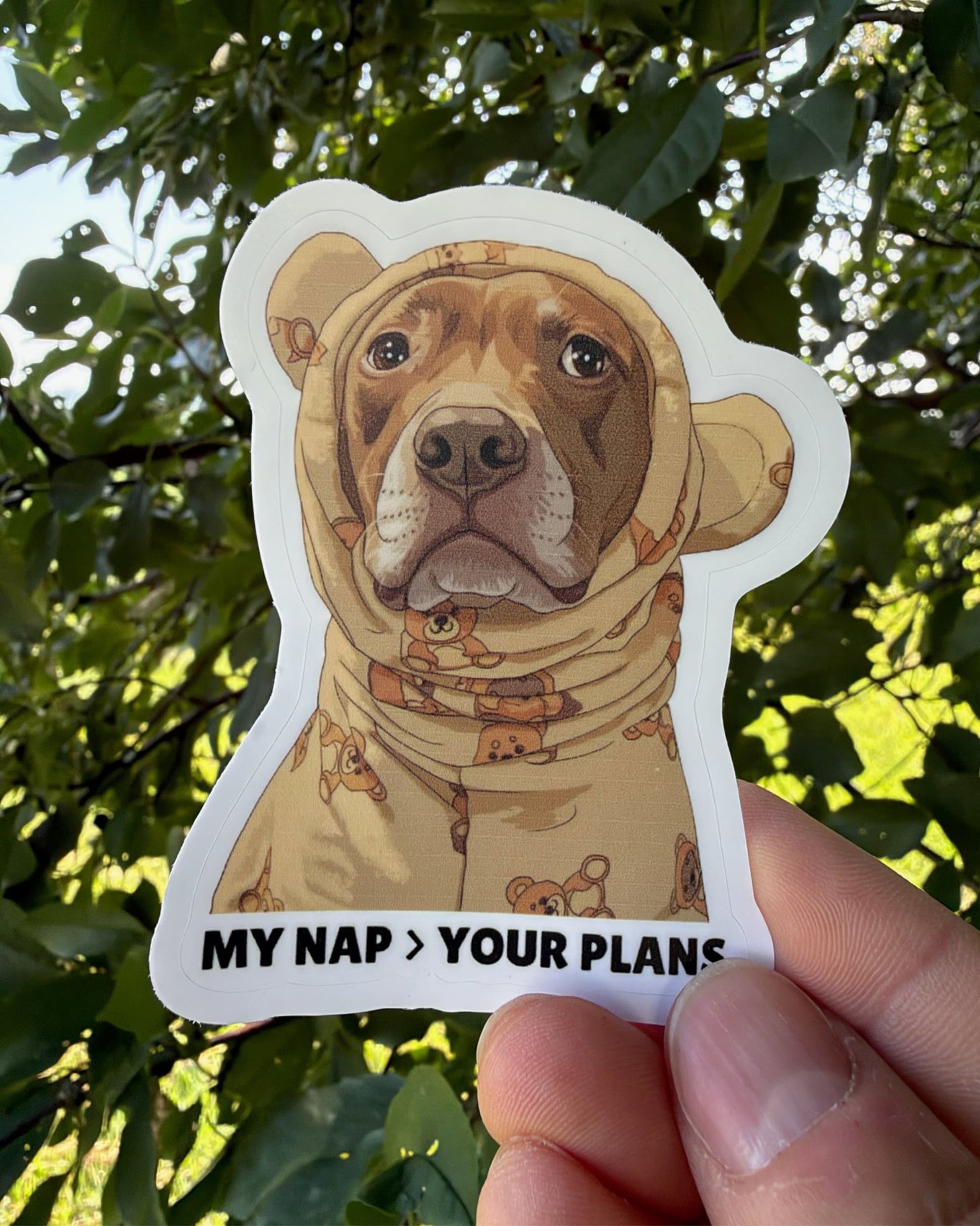 Handheld sticker in front of outdoor greenery. Opie nap sticker: showing illustration of red-brown pit bull in teddy bear pajamas kiss-cut sticker by Sly Dawg Studio. Caption at bottom: My nap > your plans