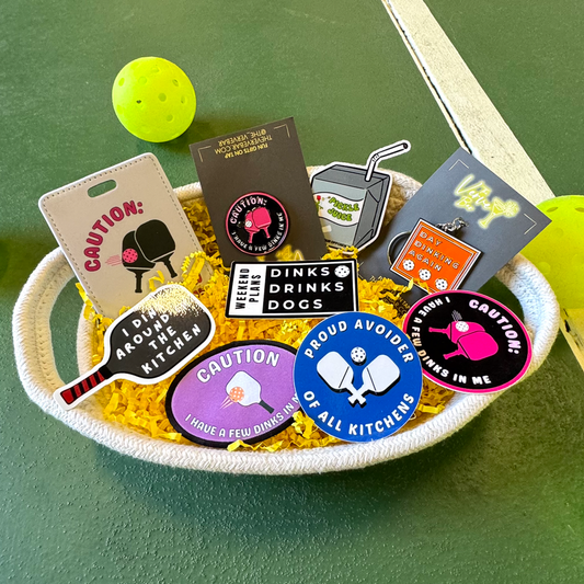 A gift basket containing various pickleball-themed items such as a luggage tag, stickers with phrases, an enamel keychain, and a rubber keychain.