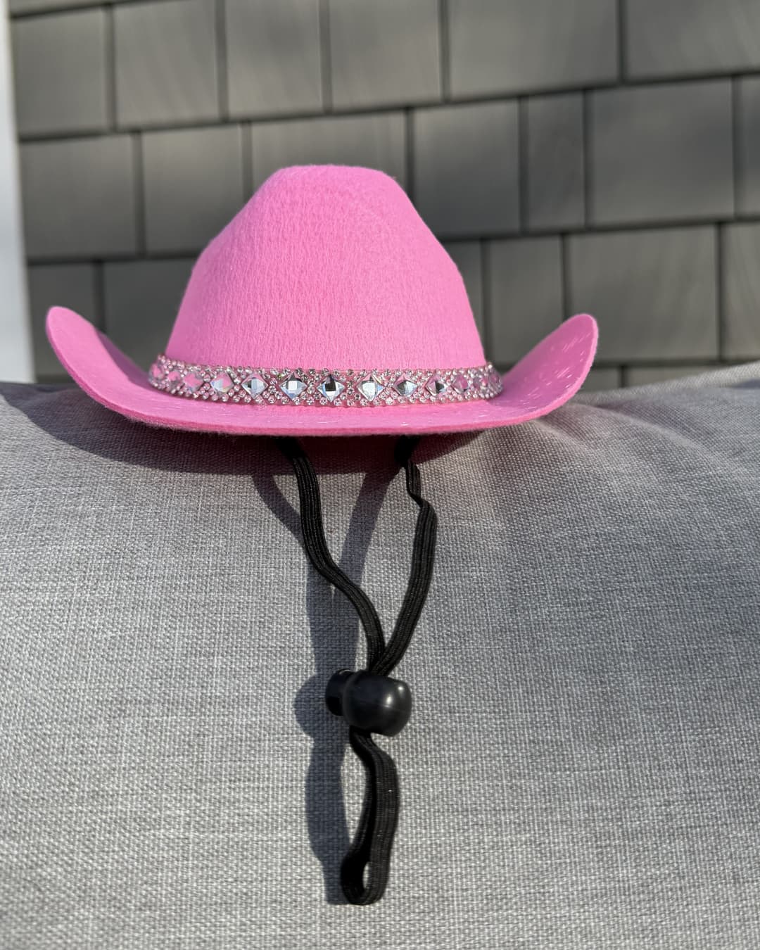 Pink cowboy hat for dogs with decorative rhinestone  band on a gray chair with hat chin strap hanging down.