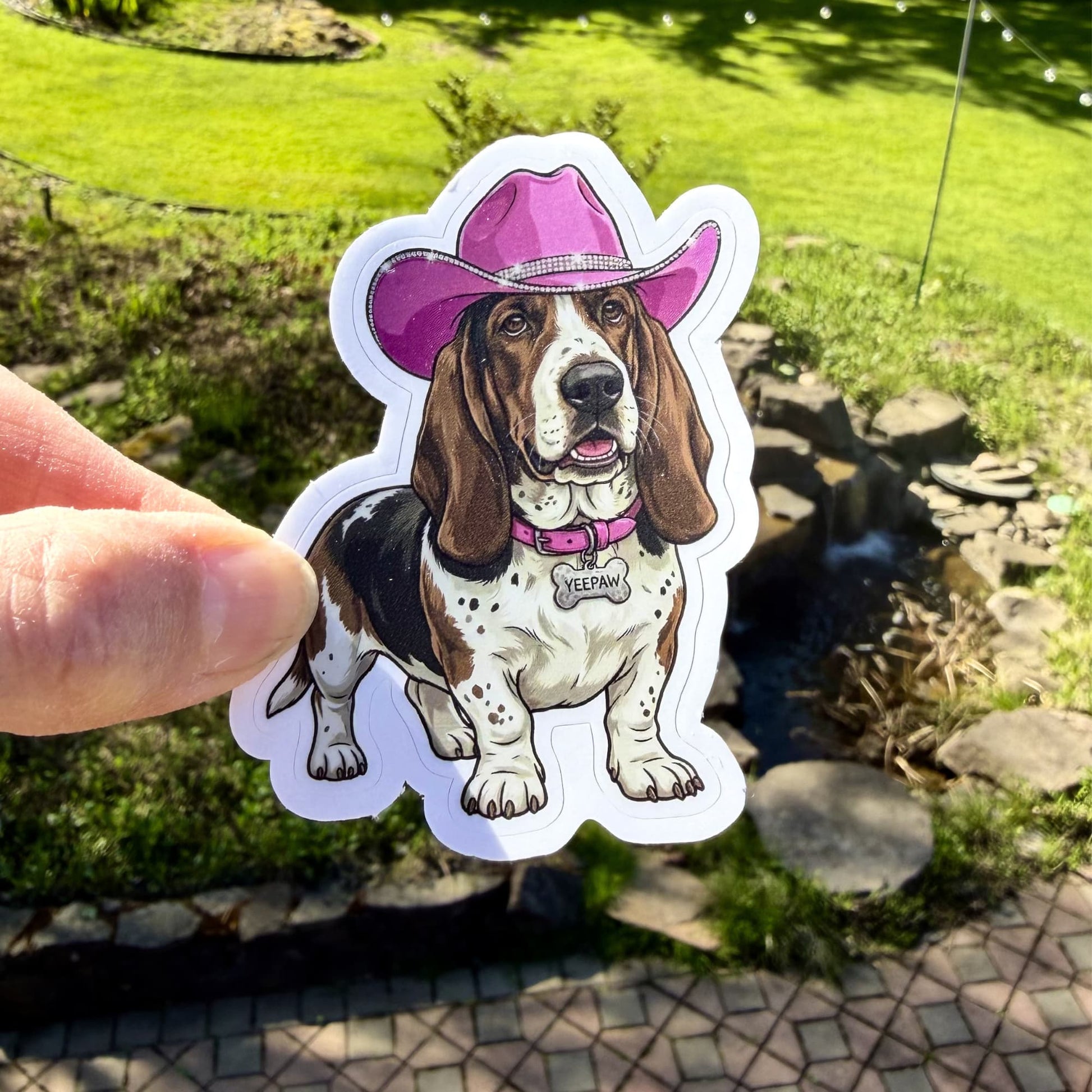 Pink cowgirl basset hound sticker; illustration of dog wearing a pink cowboy hat held by a hand outdoors.