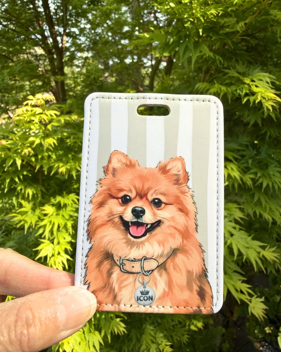Pomeranian luggage tag lifestyle; Hand holding a fun faux leather luggage tag featuring a fluffy orange Pomeranian with a 1 Brain Cell collar charm, grey and white stripes, green tree foliage background.