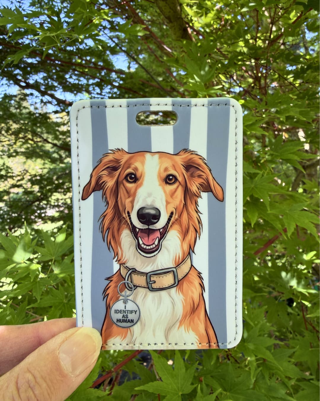 Red and white borzoi luggage tag lifestyle; Hand holding a fun faux leather luggage tag featuring an illustrated red and white Borzoi with an I Identify As Human collar charm, grey and white vertical stripes, green tree foliage background.