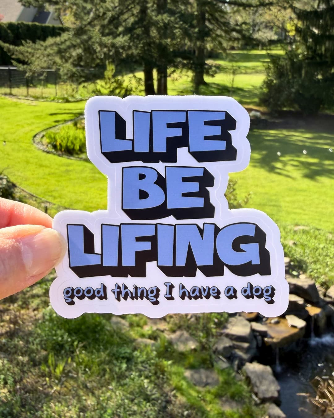 Relatable Dog Owner Sticker;  text 'LIFE BE LIVING good thing I have a dog' in bold block blue text. Hand held and nature background