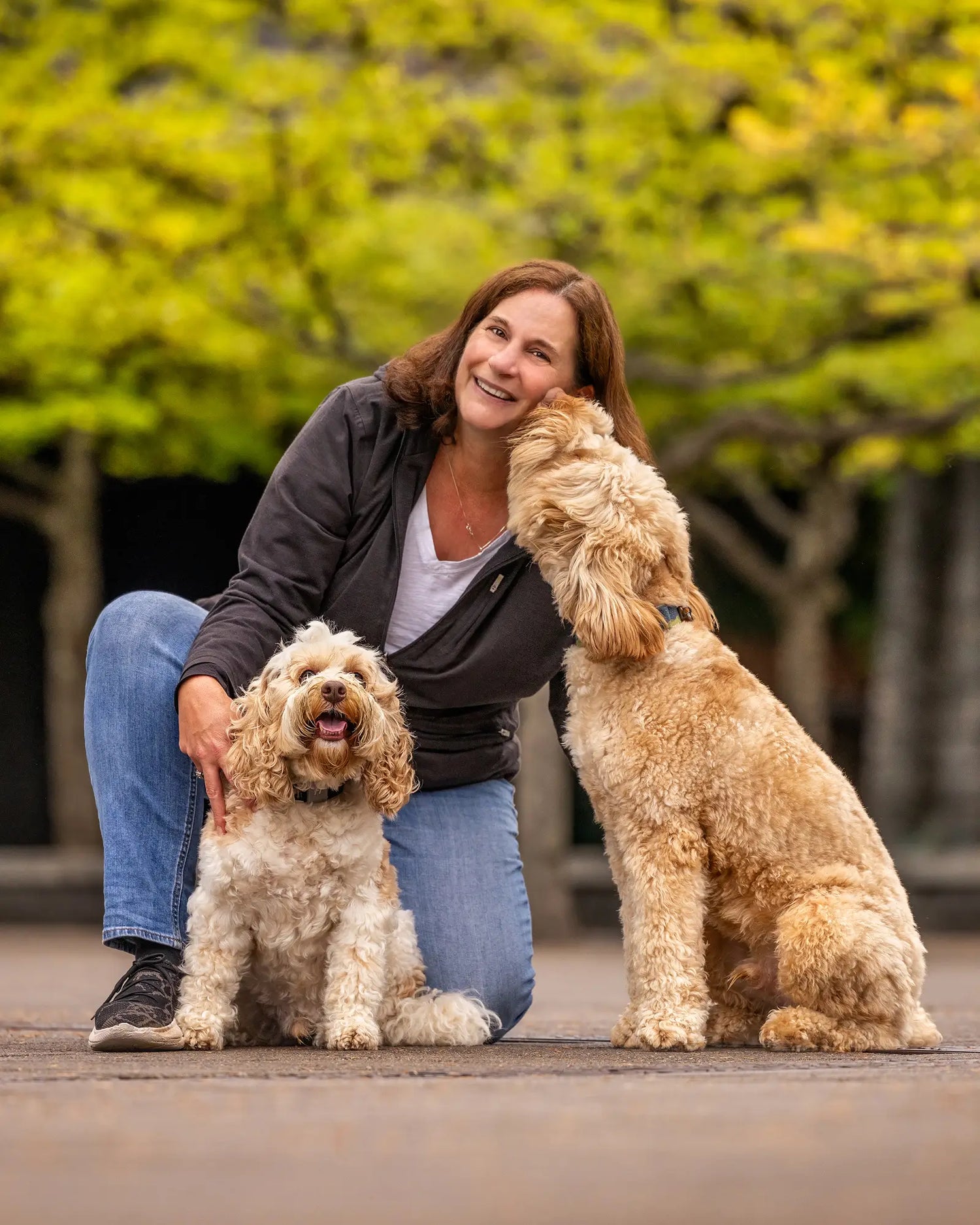Woman (owner of Sly Dawg Studio sticker shop) laughing and petting her two labradoodle dogs. Trees in background. 
