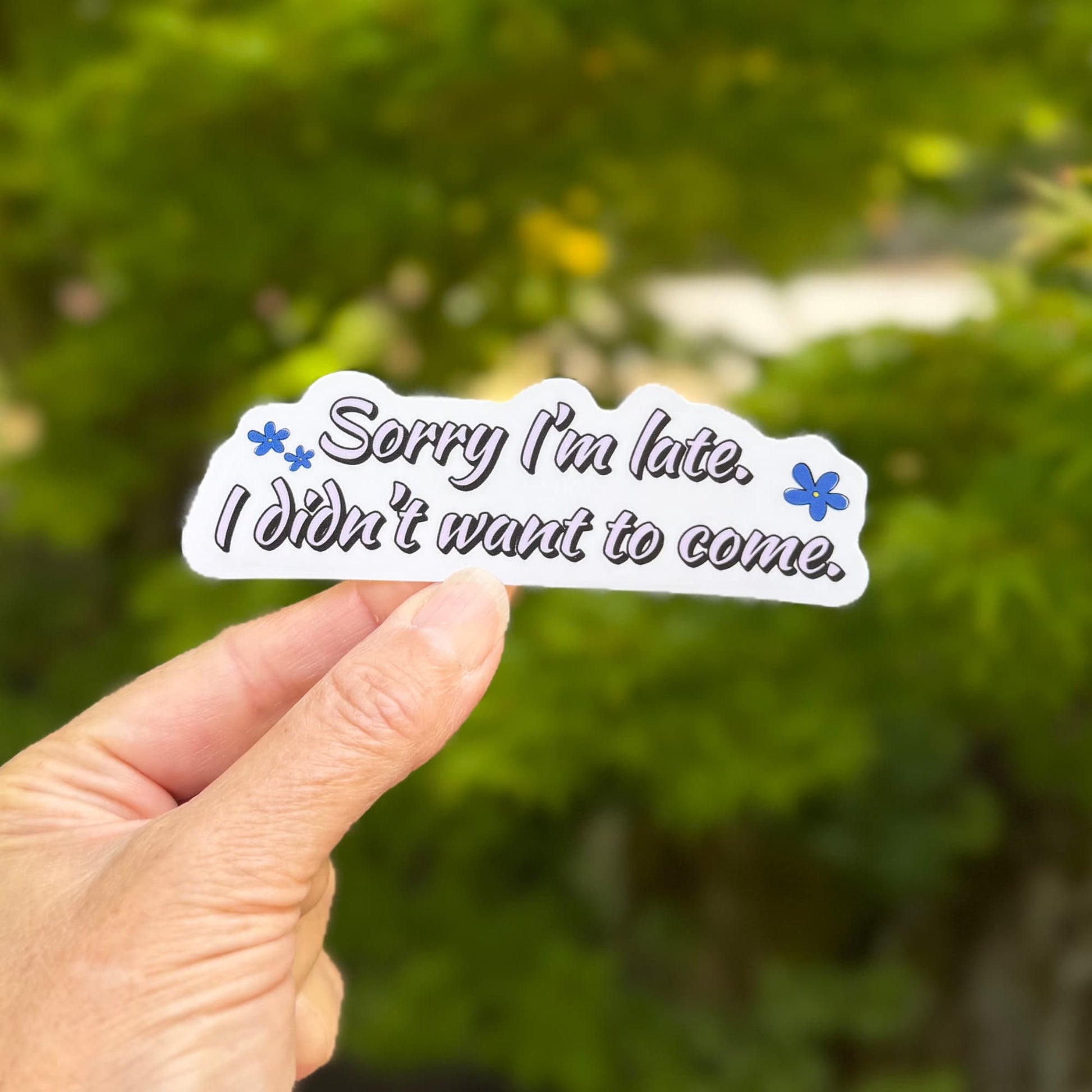 Sorry I'm late sticker held in one hand in front of leafy green trees, showing the full quote 'Sorry I'm late. I didn't want to come.' in black script