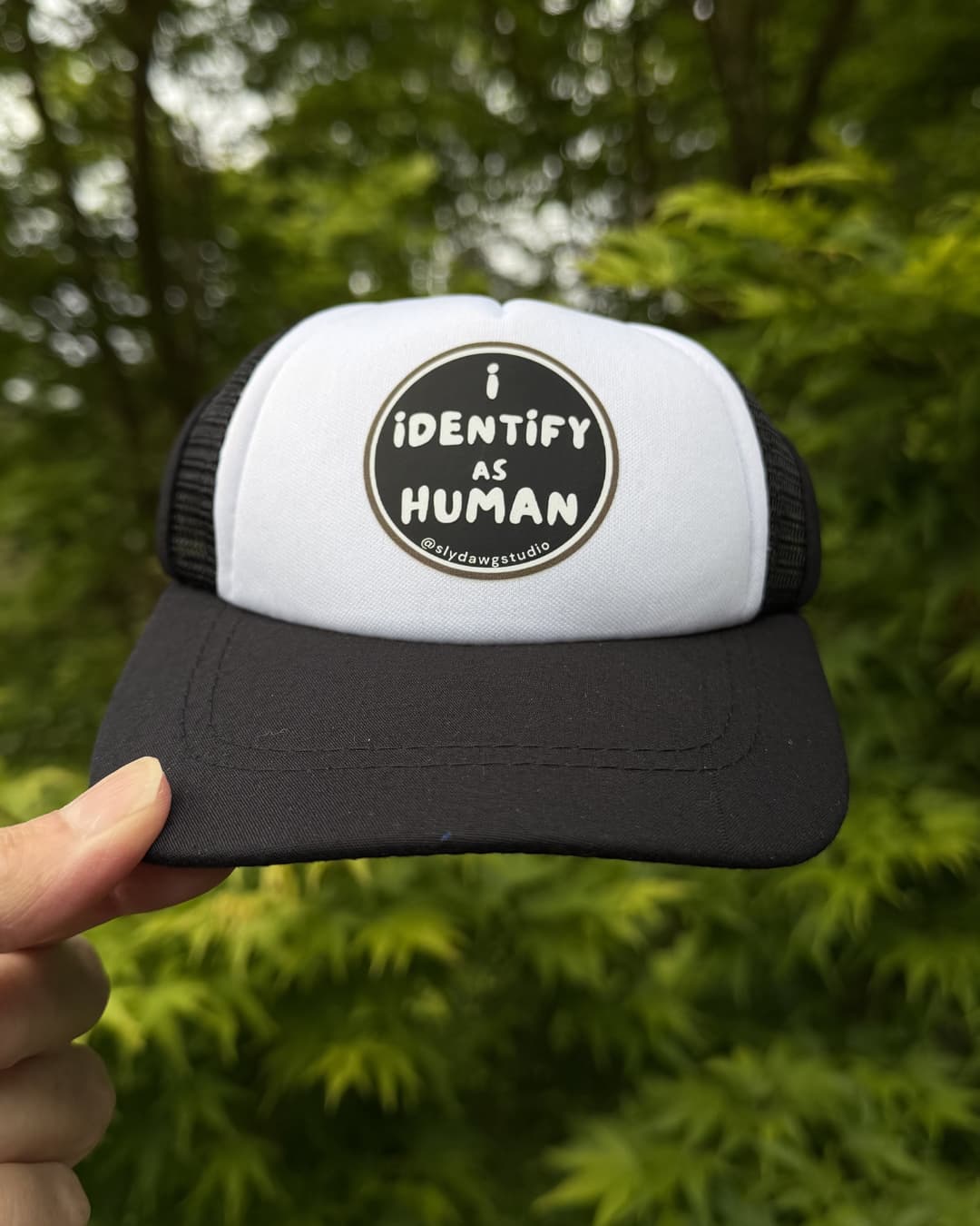 The best dog hat ever lifestyle; hand holding a black and white I Identify as Human dog trucker hat outdoors with green foliage in the background.