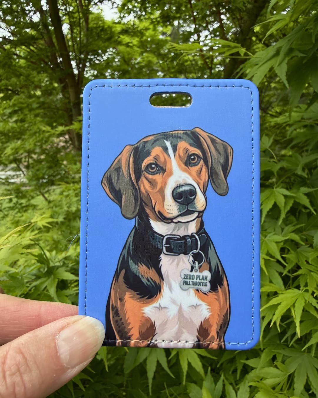 Treeing Walker Coonhound luggage tag lifestyle; Hand holding a fun faux leather luggage tag featuring a tricolor Treeing Walker Coonhound with a Zero Plan Full Throttle collar charm, solid sky blue background, green tree foliage background.
