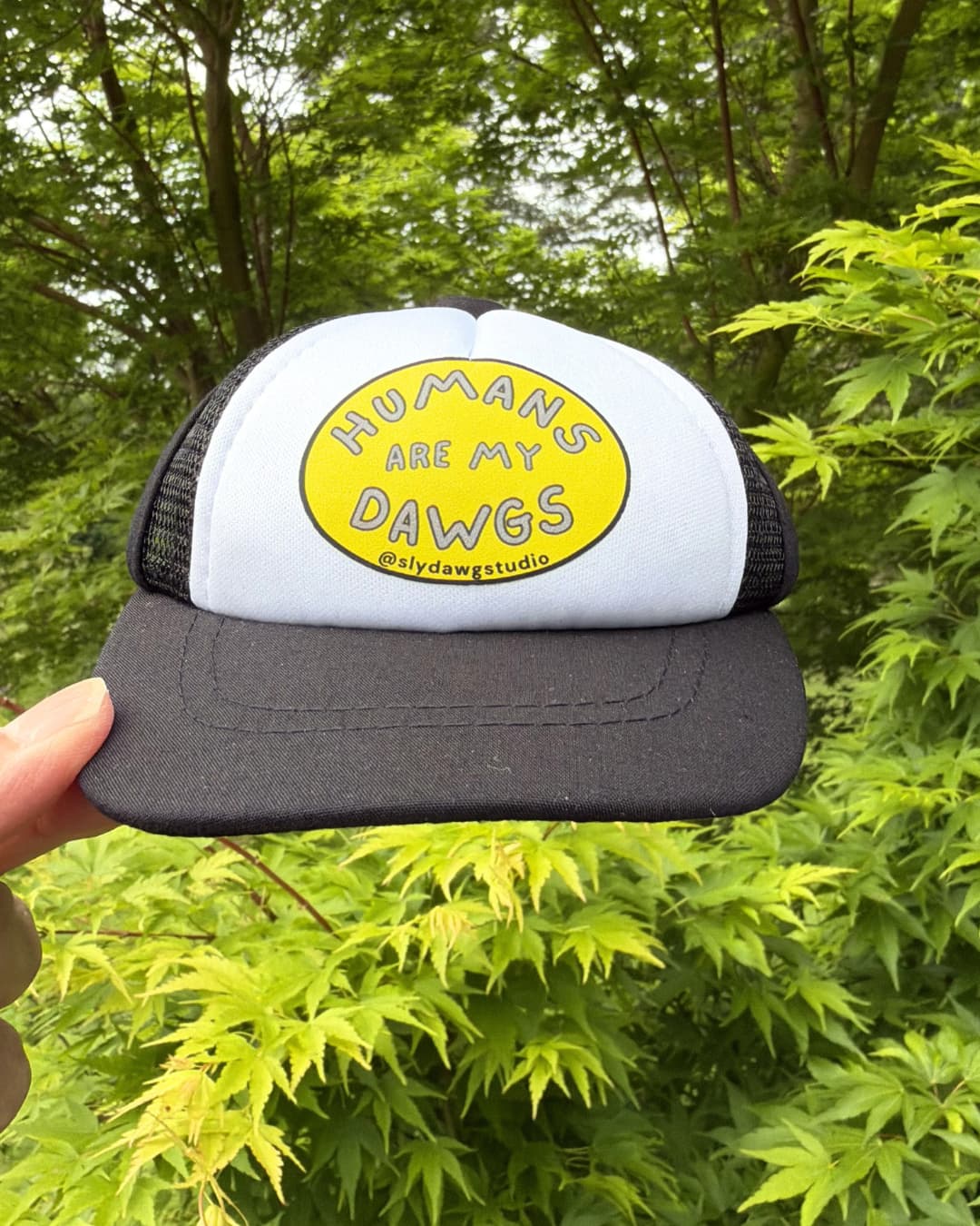 Trucker hat for dogs lifestyle; hand holding a black and white Humans Are My Dawgs dog trucker hat outdoors with green Japanese Maple leaves in the background.