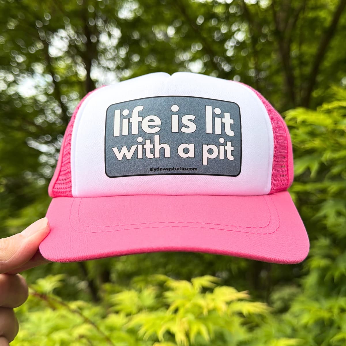 Trucker hats for pitbulls; a hand holding a pink and white Life is Lit with a Pit trucker hat outdoors with green leaves in the background.