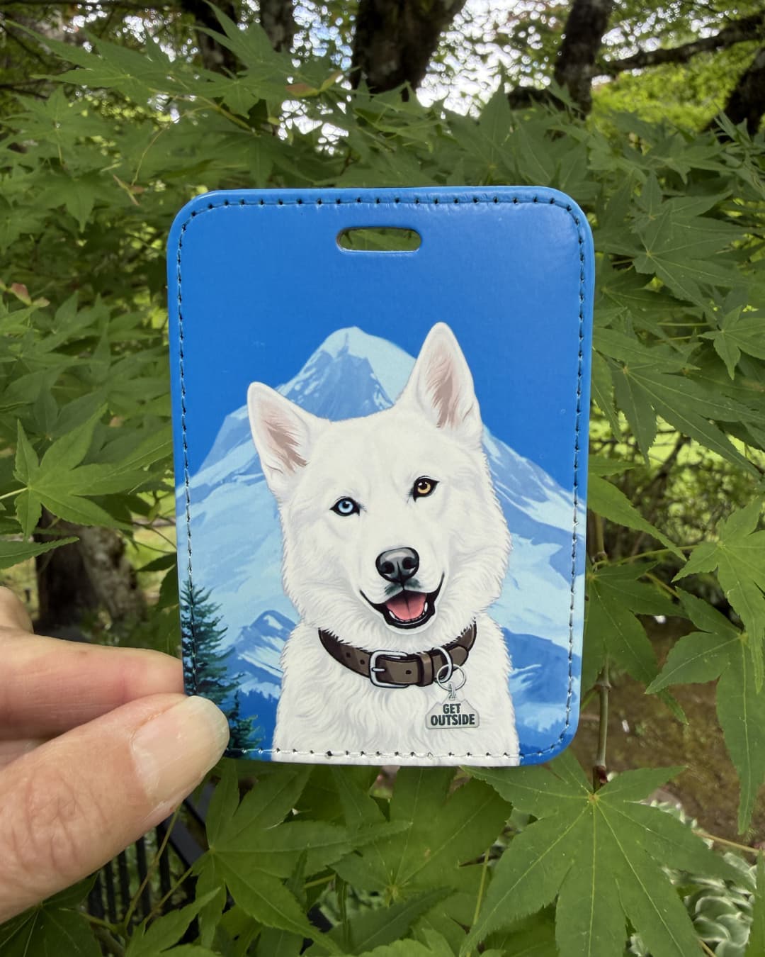 White husky dog luggage tag lifestyle; Hand holding a fun faux leather luggage tag featuring a pure white Siberian Husky with heterochromia, dark brown collar with Get Outside charm, vivid blue sky and mountain background, green foliage surroundings.