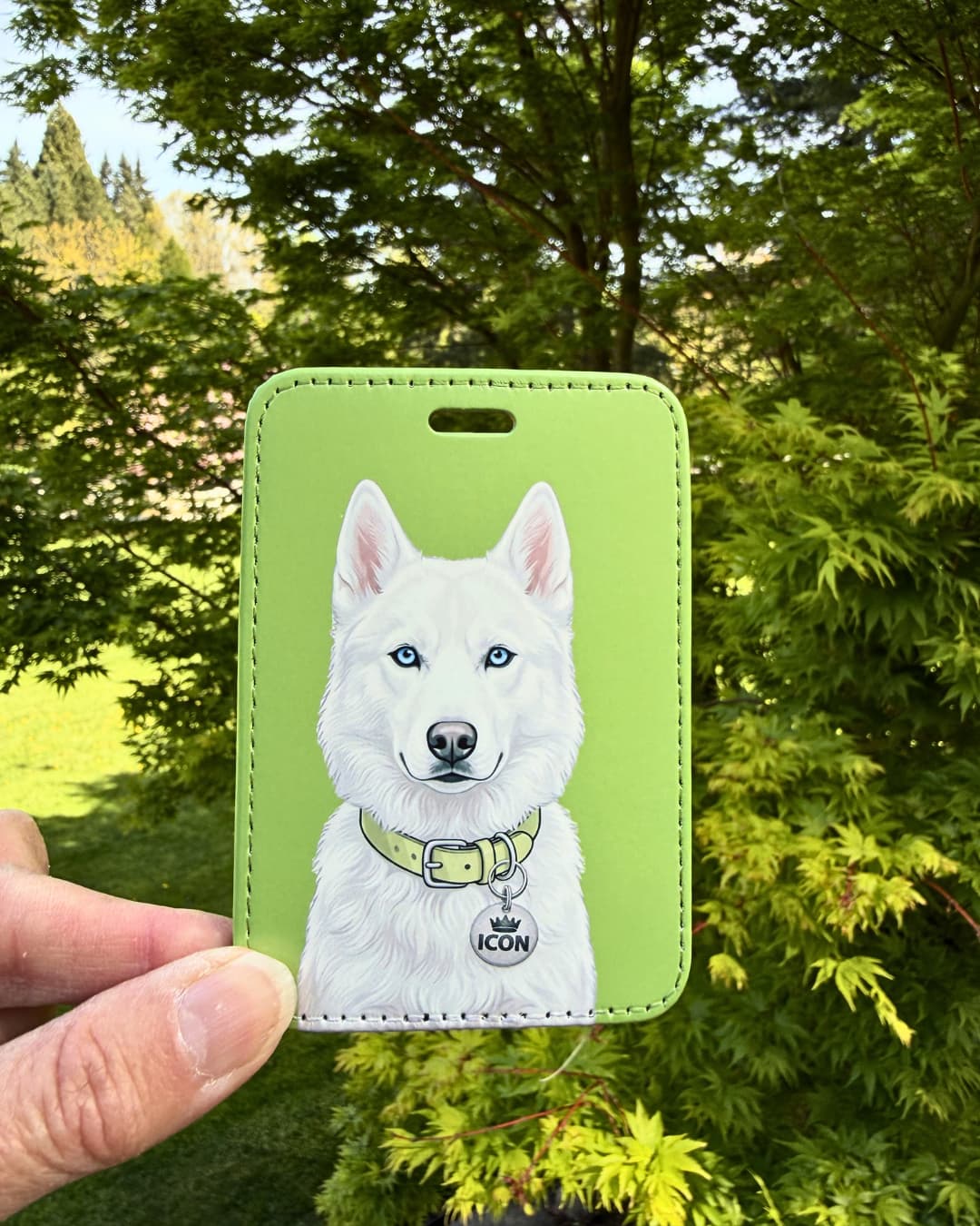 white husky luggage tag held outdoors against green leaf background showing detailed white fur and blue eye illustration with ICON collar tag